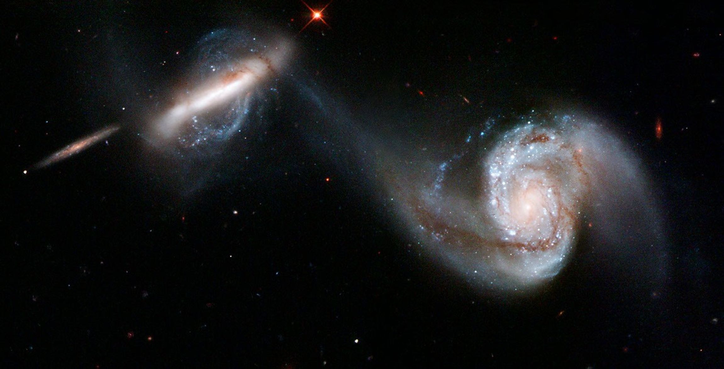 A pair of merging galaxies. The galaxy on the right has a large, single spiral arm curving out from the core and around to above it, with very visible glowing dust and gas. The left galaxy is shaped like a long bar., surrounded by swirls of blue-white stars. A thin stream of whitish gas connects the two galaxies' cores. A few small stars and galaxies are scattered around the black background.